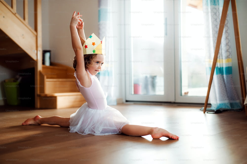 家に王女の王冠を持つ小さなバレリーナの女の子で、体操のスプリットをしています。