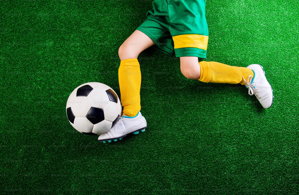 Legs of unrecognizable little football player with soccer ball against artificial grass. Studio shot on green grass.