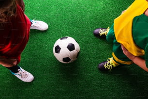 Piernas de dos pequeños jugadores de fútbol irreconocibles con balón de fútbol contra césped artificial. Estudio filmado sobre fondo verde.