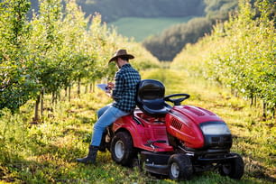 Un agriculteur d’âge mûr debout à l’extérieur par un mini tracteur dans un verger, utilisant une tablette.