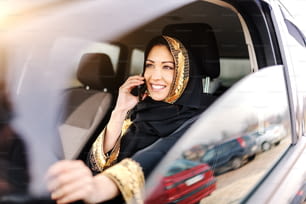 Bella donna musulmana con sorriso dentato e sciarpa in testa che parla al telefono e tiene la mano sul volante.