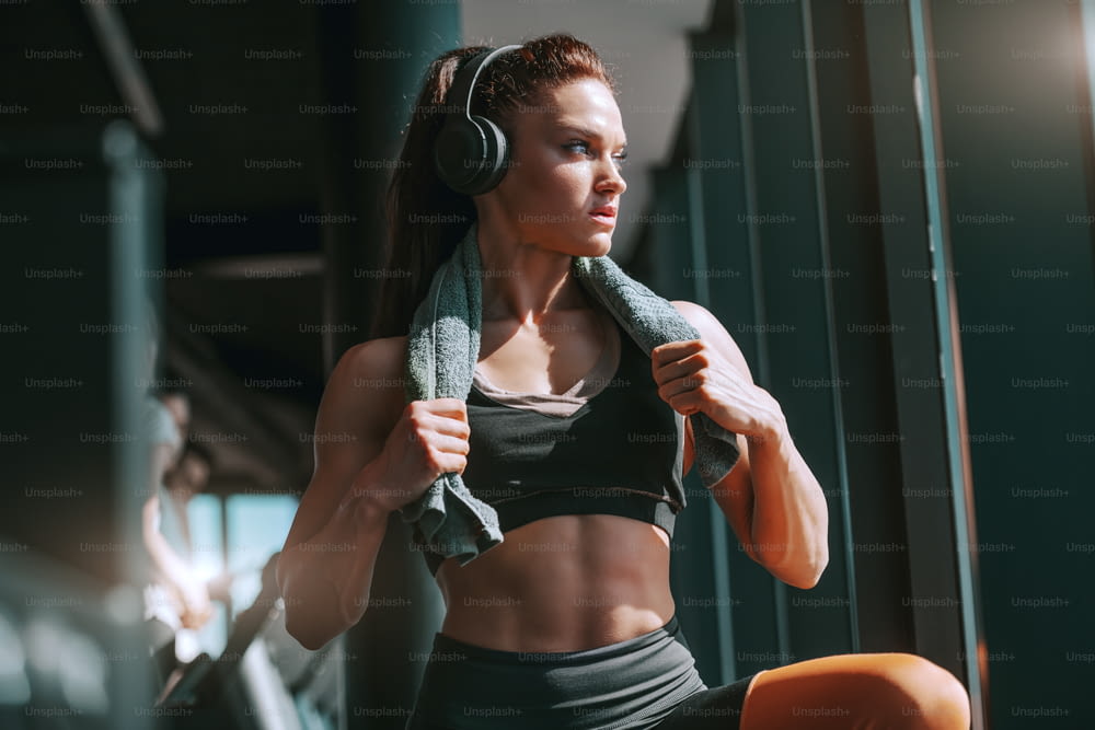 Young attractive strong muscular female bodybuilder with ponytail and headphones posing in gym with towel around neck. Dreams don't work unless you do.