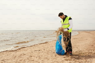 Young volunteer in protective workwear standing on sandy beach while putting natural wastes into blue sack during work