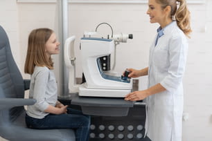 Retrato de vista lateral de un oftalmólogo amigable usando un refractómetro automático mientras examina los ojos del niño. Ellos mirando a cada uno y sonriendo
