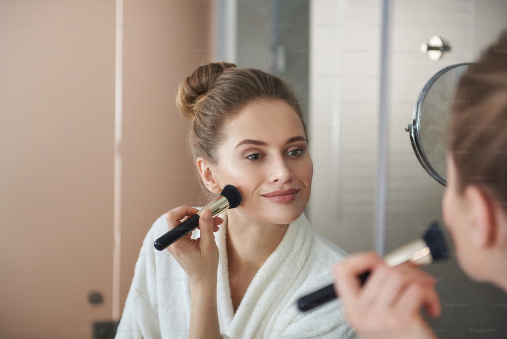 Autocuidado e conceito de maquiagem. Close up reflexo no espelho da mulher jovem sorridente no roupão branco deitado em pó blusher usando escova
