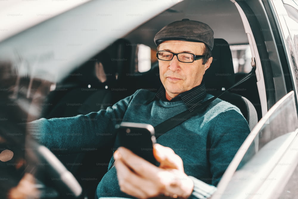 Serious Caucasian senior man with cap on head and eyeglasses sitting in car and using smart phone. Window opened.