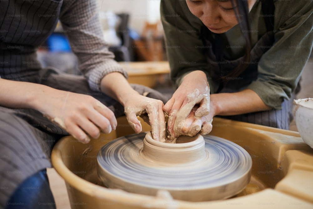 Close-up of skilled Asian female potter showing woman how to mould clay dish using pottery wheel in workshop