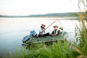 Grandfather with adult son fishing on the inflatable boat on the lake with green cane on the foreground early in the morning