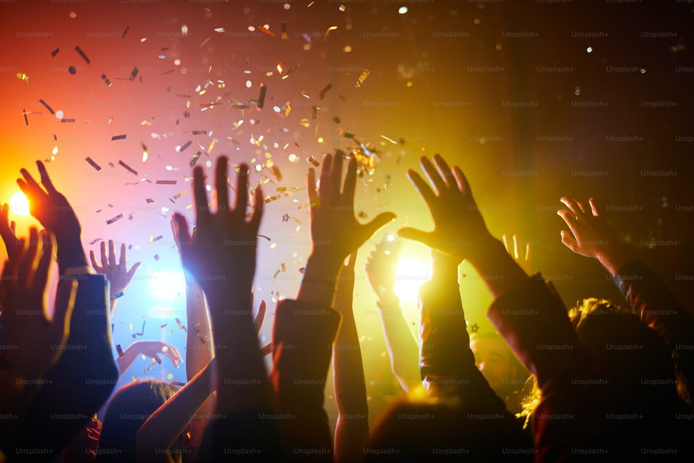 Groupe de personnes méconnaissables debout sous des confettis et levant les mains en l’air lors d’un spectacle musical