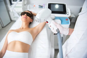 Aesthetic body treatment. Close up top angle portrait of cosmetologist holding laser hair removal device in hand while young woman in eyeglasses lying on couch
