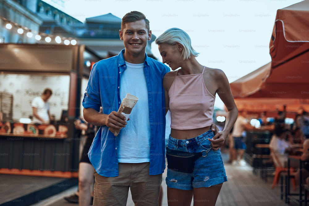 Positive man hugging girlfriend at the fair and smiling while holding doner kebab