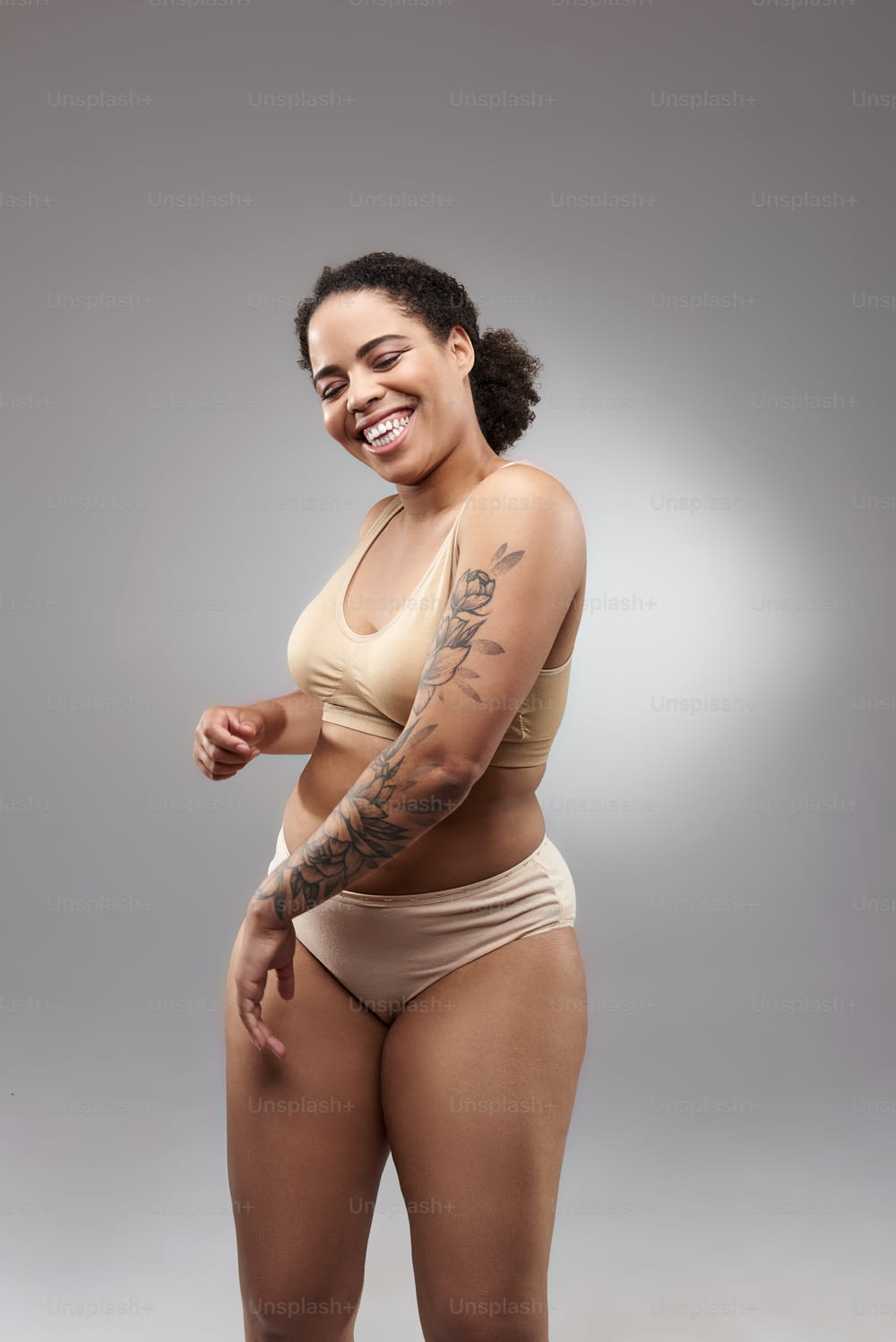 Smiling lady with curly hair wearing light underwear and showing her tattoo. Overweight concept