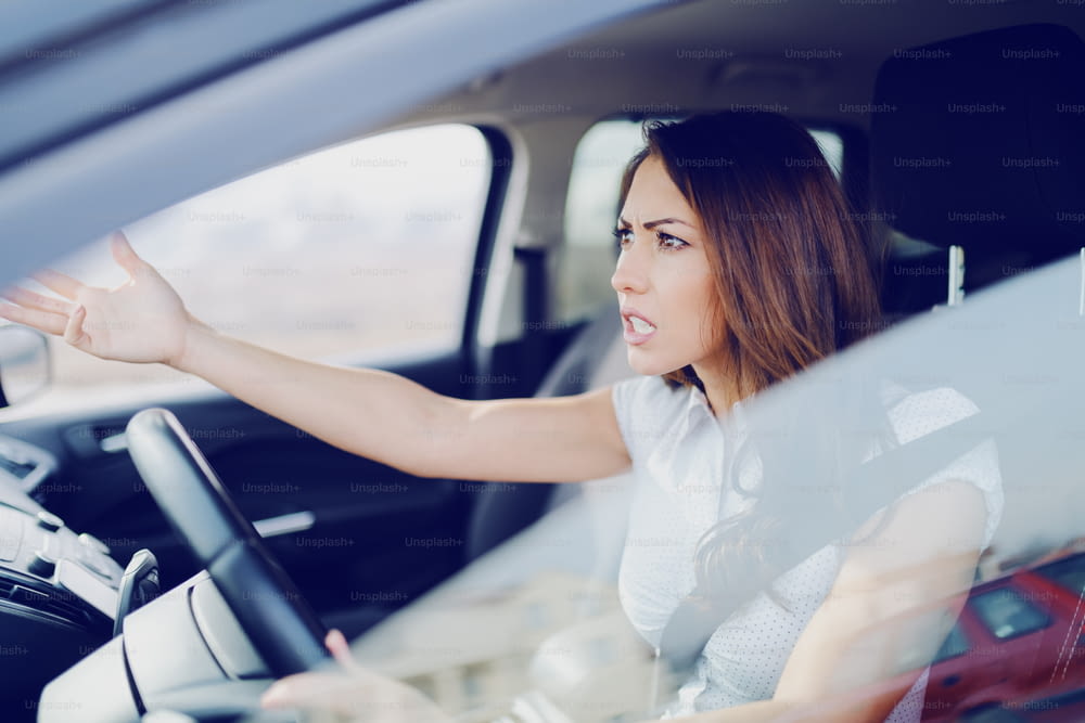 Angry attractive caucasian brunette yelling at other drivers while sitting in car.