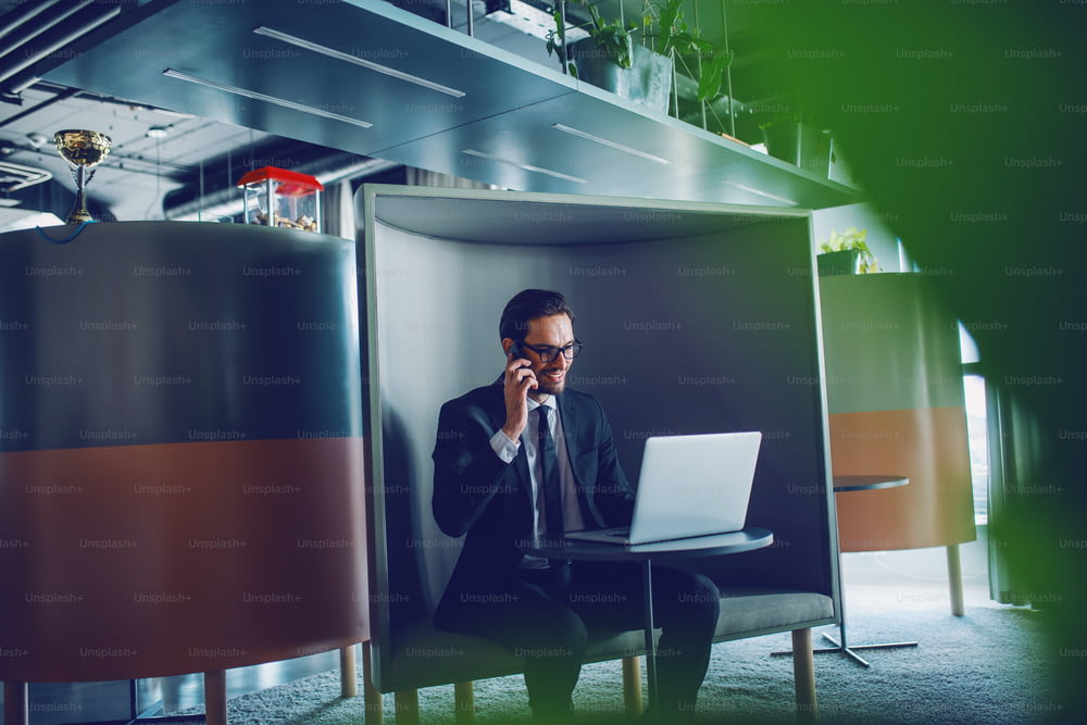 Empresário branco bonito sorridente de terno e com óculos sentado no local de trabalho, tendo chamada de negócios e olhando para laptop.