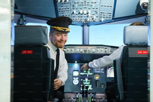 Ritratto di sorridente bel pilota barbuto in berretto da capitano seduto in poltrona di pelle e voltandosi indietro nella cabina di pilotaggio
