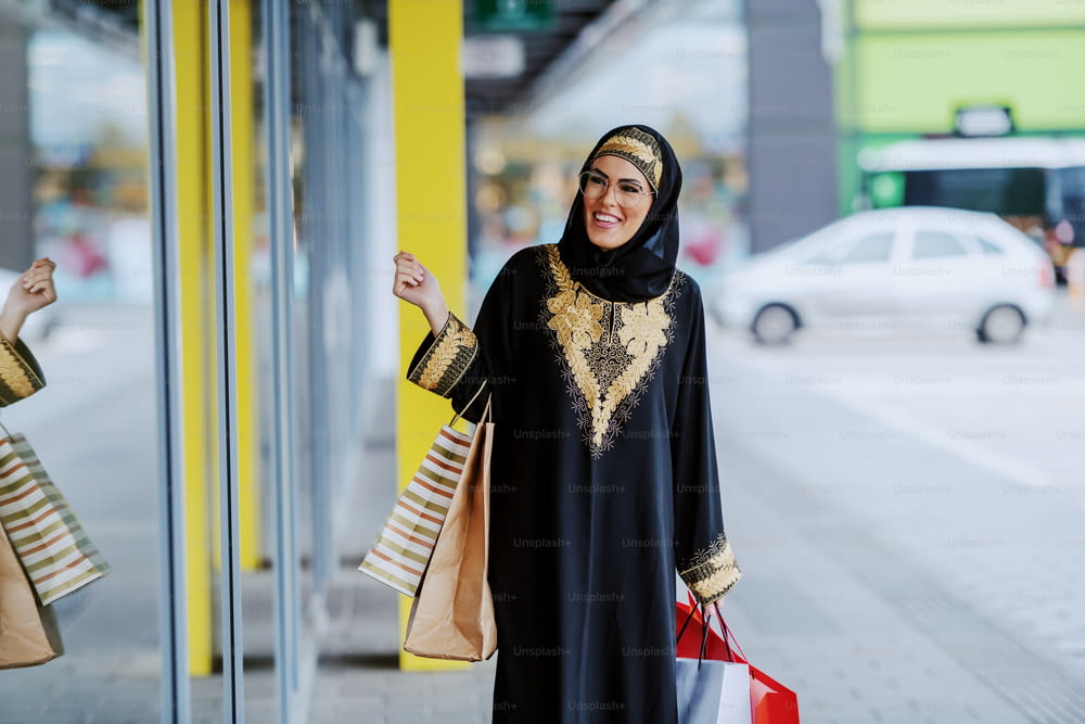 Attractive smiling arab woman in traditional wear looking at shop window while standing with shopping bags in hands.