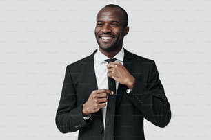Eleganter junger afrikanischer Mann in formeller Kleidung, der die Krawatte zurechtrückt und lächelt, während er vor grauem Hintergrund steht