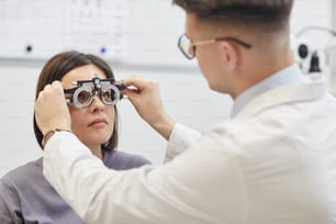 Retrato de un joven oftalmólogo que pone marco de prueba en una paciente durante el control de la vista en una clínica moderna, espacio de copia