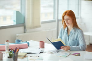 Porträt einer sommersprossigen jungen Frau, die Tagebuch liest, während sie am kreativen Arbeitsplatz am Fenster sitzt, Kopierraum