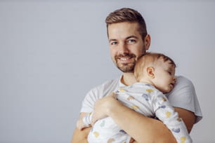 Un jeune père souriant et séduisant et non rasé a finalement réussi à endormir son fils bien-aimé. Le père tient son enfant dans ses bras et regarde la caméra.