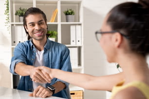 Happy young successful businessman shaking hand of new business partner after making deal while looking at her with smile