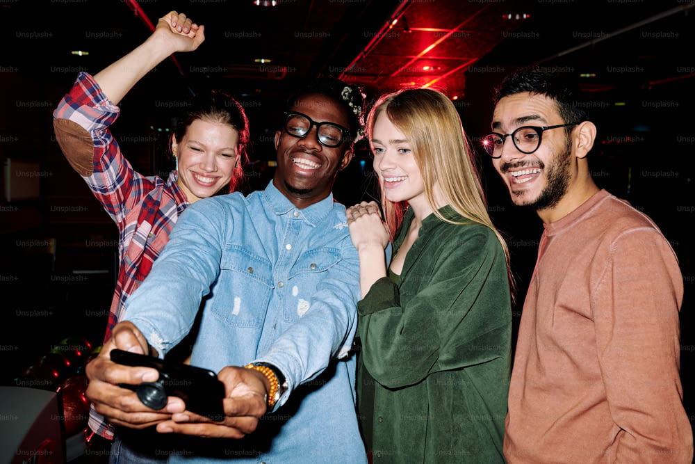 Quattro giovani uomini e donne gioiosi in abbigliamento casual che guardano la fotocamera dello smartphone e fanno selfie mentre si godono la festa in club