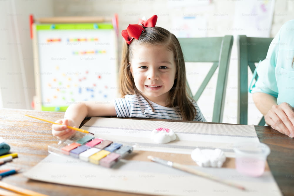 Nette kleine Dreijährige, die Aquarellfarben verwendet, um eine Gips-Cupcake-Figur zu malen und mit ihrer Mutter die Schule zu Hause zu genießen