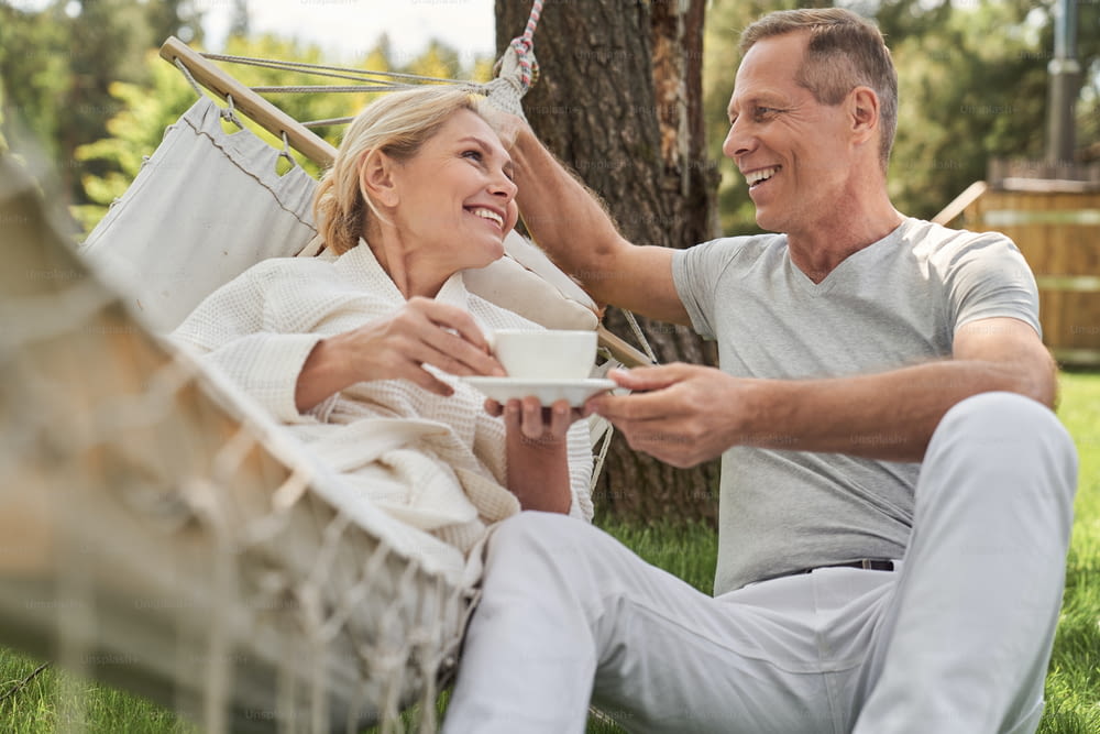 Happy woman with cup of tea lying in hammock and talking to husband in open air