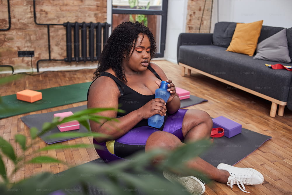 Curly calm lady keeping a bottle with water and sitting in crossed position while having a rest after doing exercises in living room