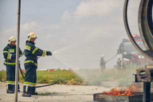 Seitenansicht von zwei Feuerwehrleuten in Schutzuniform und gelben Helmen, die Wasser aus dem Schlauch spritzen