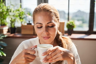 Calm smiling woman in a bathrobe sitting with a white cup of coffee and looking at it