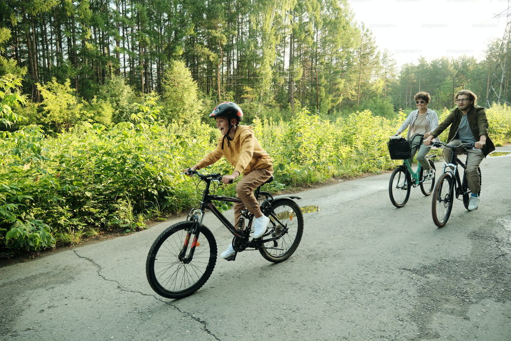 Young couple and their son riding bicycles along road in natural environment against green trees and bushes on summer weekend