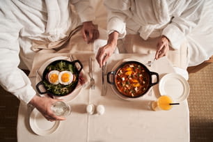 Couple in bathrobes sitting at the table with egg meals and healthy beverages. Top view of the table with food