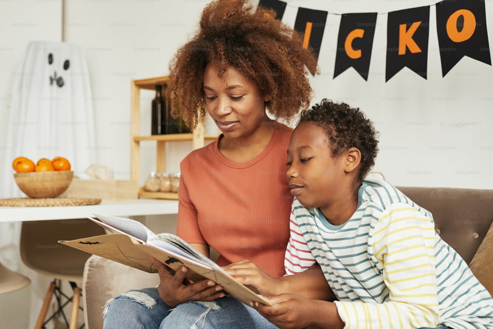 Schöne afroamerikanische Frau, die mit ihrem jugendlichen Sohn auf dem Sofa im Wohnzimmer sitzt und interessante Halloween-Geschichten liest