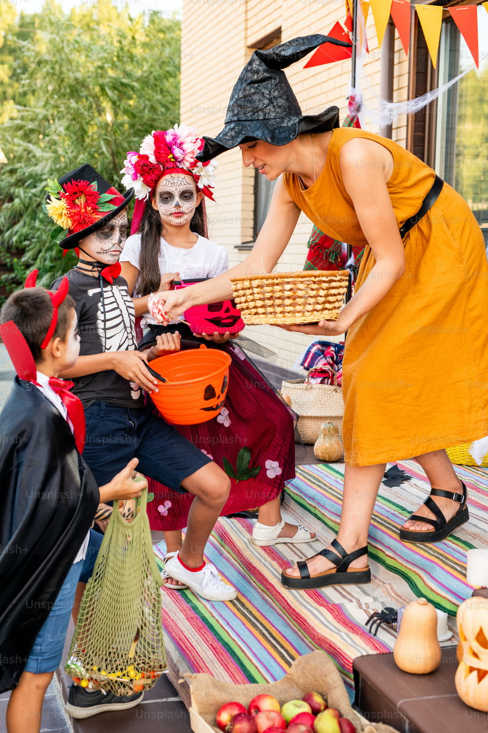 Young female in yellow dress and black witch hat bending in front of group of spooky halloween kids while giving them sweet treats from basket
