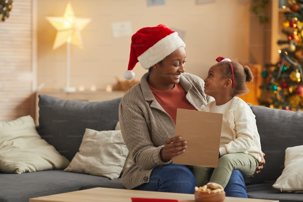 Portrait of smiling African-American mother reading letter to Santa with cute little girl while enjoying Christmas season at home, copy space