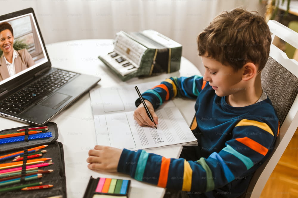 Little boy writes musical notes and violin key at home online course on laptop.Online training, online classes.
