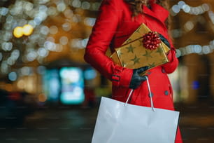 冬の楽しみ。夕方、街の屋外で買い物袋やギフトを持つ赤いコートと黒いベレー帽を着た中年の旅行者女性の接写。