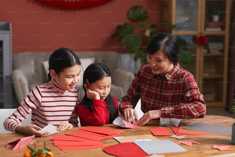 Chinesin sitzt am Tisch im Wohnzimmer und zeigt ihren kleinen Töchtern, wie man Papierkraniche herstellt
