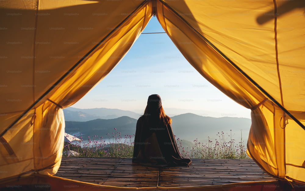 Rückansicht einer Frau, die auf einem Holzbalkon sitzt und einen wunderschönen Blick auf die Berge und die Natur außerhalb des Zeltes beobachtet