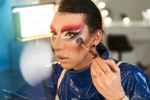 Portrait view of the drag queen with bright makeup applying highlighter at his face while sitting at the dressing table. âDrag queen and transgender person concept