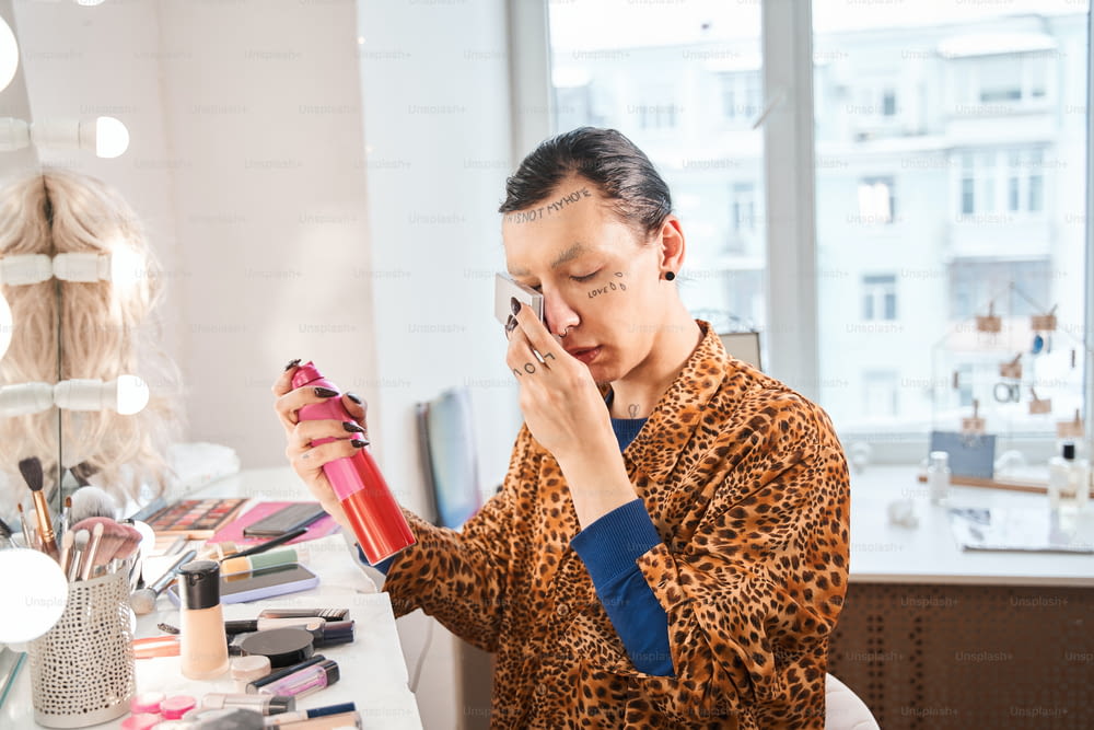 Preparing makeup. Waist up portrait view of the drag queen sitting in front of the mirror and spraying hair spray while getting ready to his new look. Drag queen and transgender person concept. Stock photo