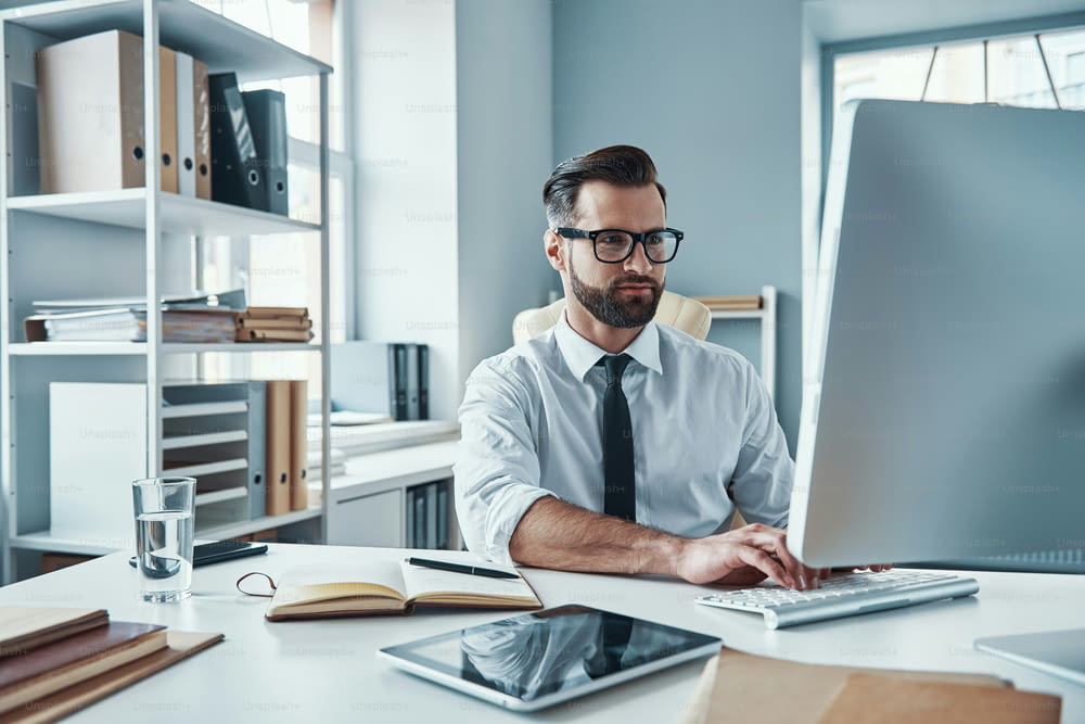 Moderner junger Mann in formeller Kleidung, der mit Technologien arbeitet, während er im Büro sitzt