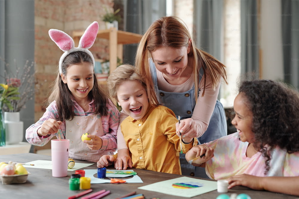 Young cheerful woman bending over table with group of happy kids while helping them with painting picture of Easter egg before holiday