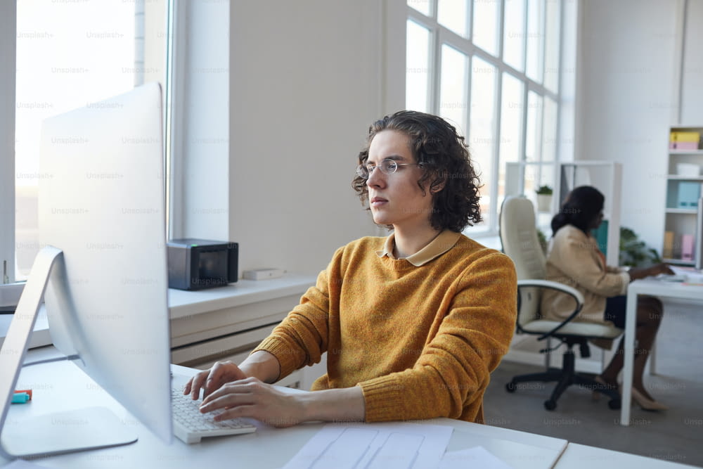 Retrato da vista lateral do jovem desenvolvedor de software usando o computador enquanto trabalha na mesa no interior branco do escritório, espaço de cópia