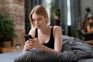 Giovane donna bionda che usa il telefono cellulare nel suo lavoro online mentre è seduta a letto al mattino