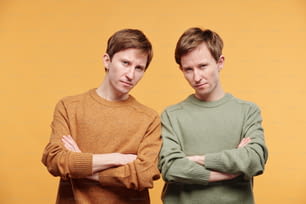 Portrait of frowning tween brothers in mustard and olive sweaters standing with crossed arms against orange background