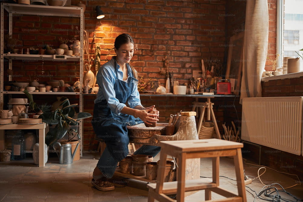 Retrato de vista lateral de cuerpo entero de una hermosa artesana enfocada sentada en un estudio de arte mientras da forma al producto cerámico de la arcilla fresca