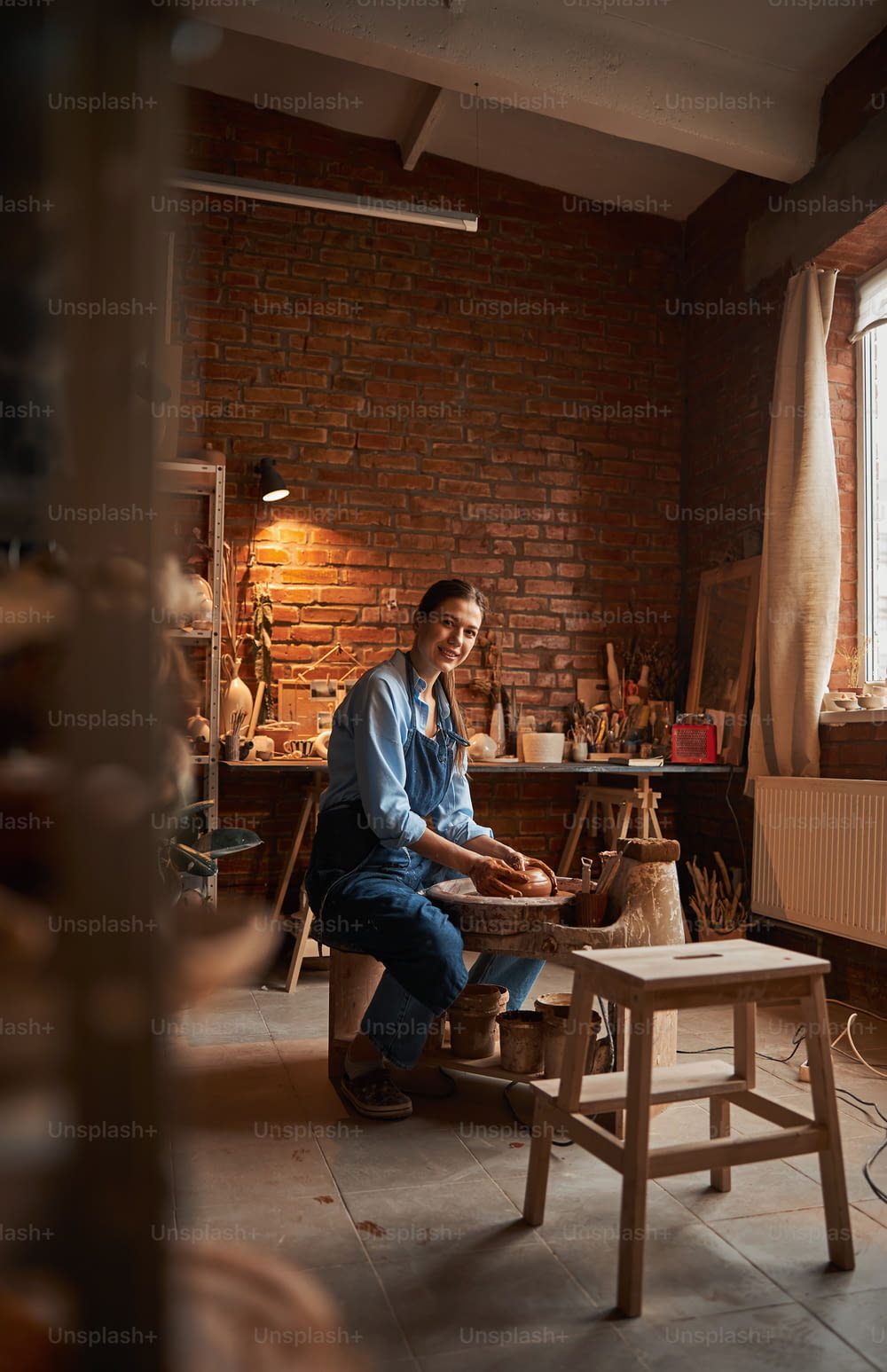 Full length side view portrait of beautiful elegant craft woman sitting near the pottery wheel while making something in art studio