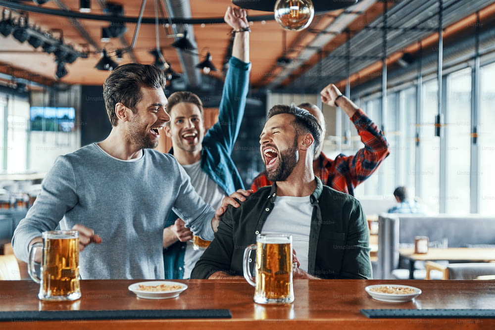 Giovani uomini eccitati in abbigliamento casual che bevono birra e guardano giochi sportivi seduti al pub